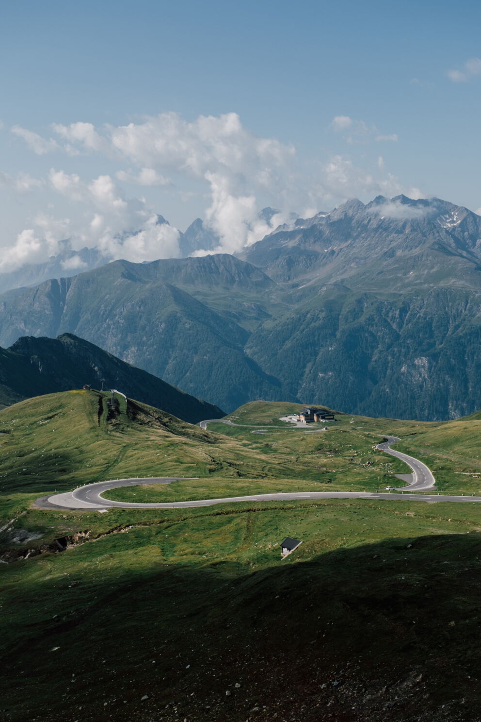 Winding mountain road through green hills in front of an impressive mountain panorama and blue sky.
