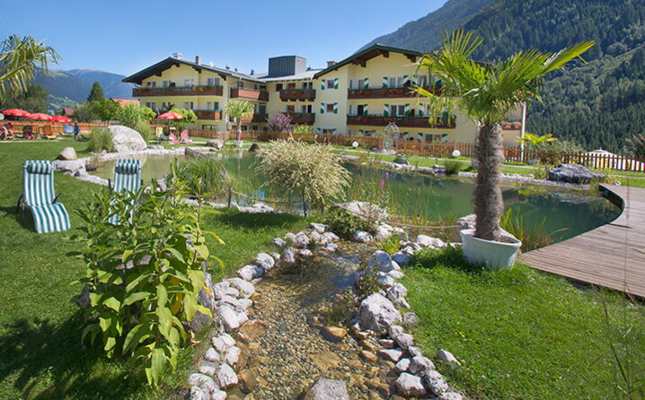 Hotel with green sunbathing lawn, palm trees and bio-pond against an alpine mountain backdrop.