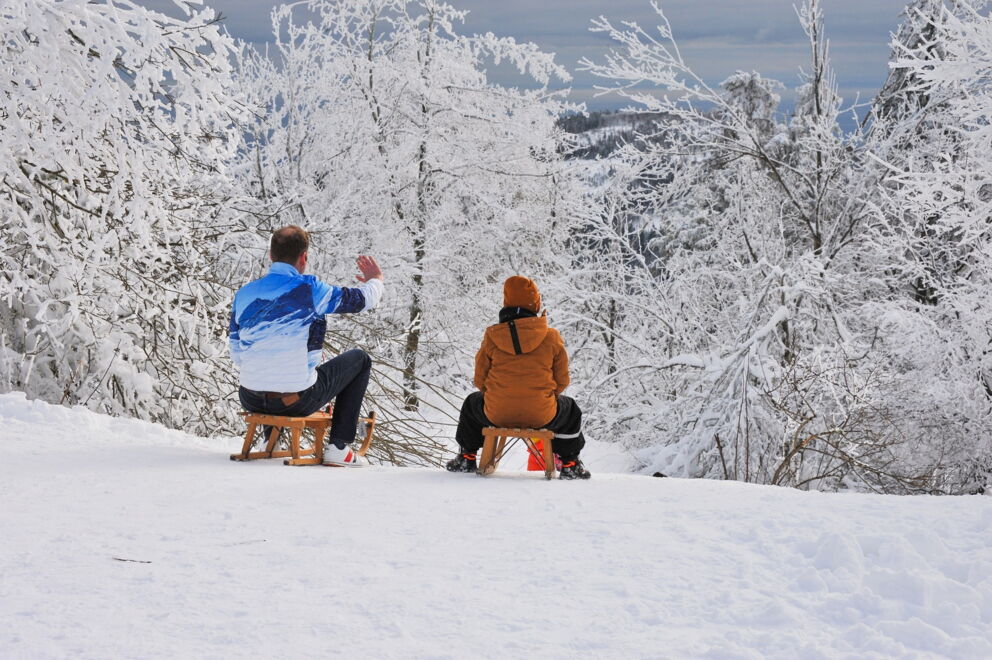 Zwei Personen rodeln im verschneiten Winterwald mit Blick auf die Berge.
