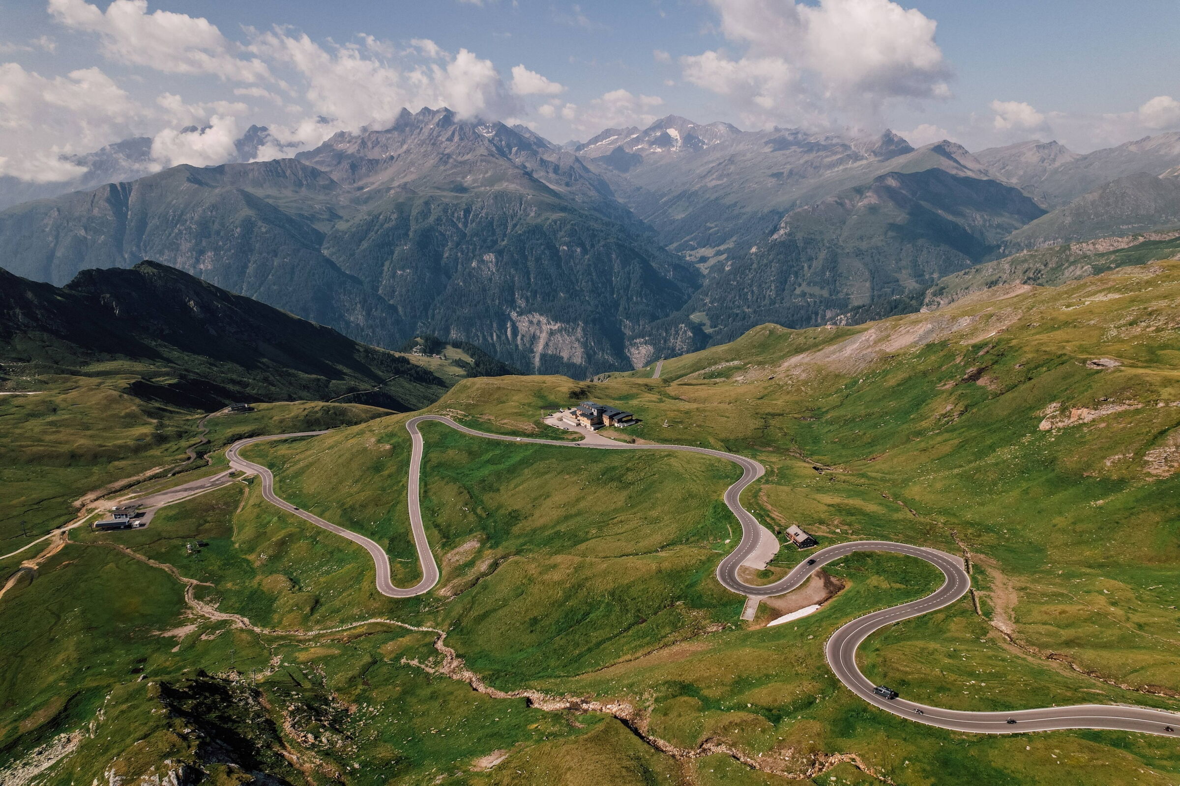 Geschwungene Straße durch grüne Alpwiesen mit majestätischen Berggipfeln im Hintergrund.