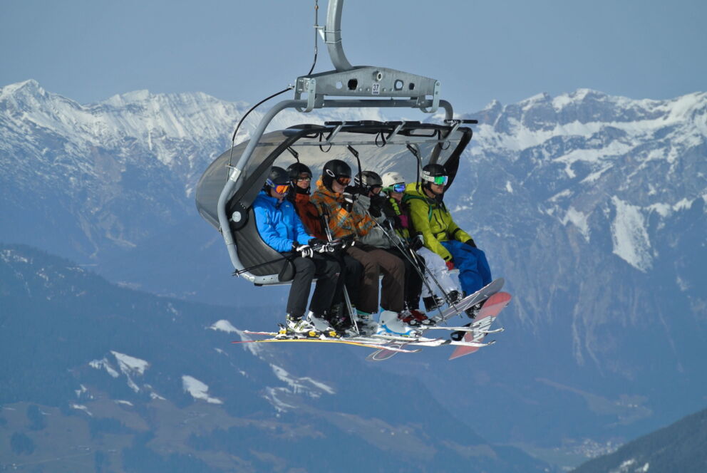 Skifahrer auf Sessellift mit beeindruckendem Bergpanorama im Hintergrund.