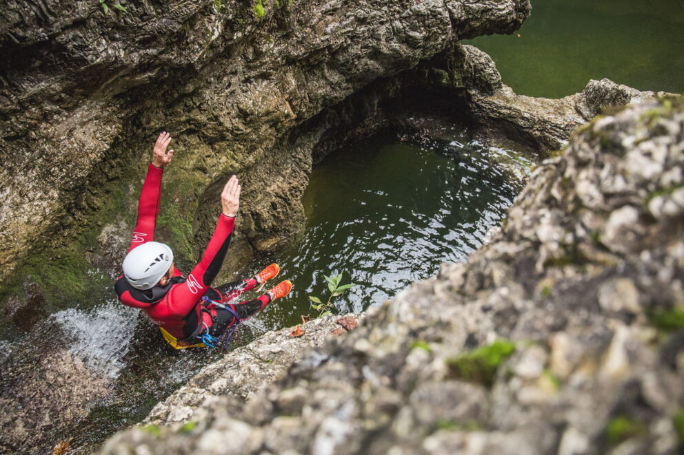 Person im Neoprenanzug springt von Felsen in klares Wasserbecken