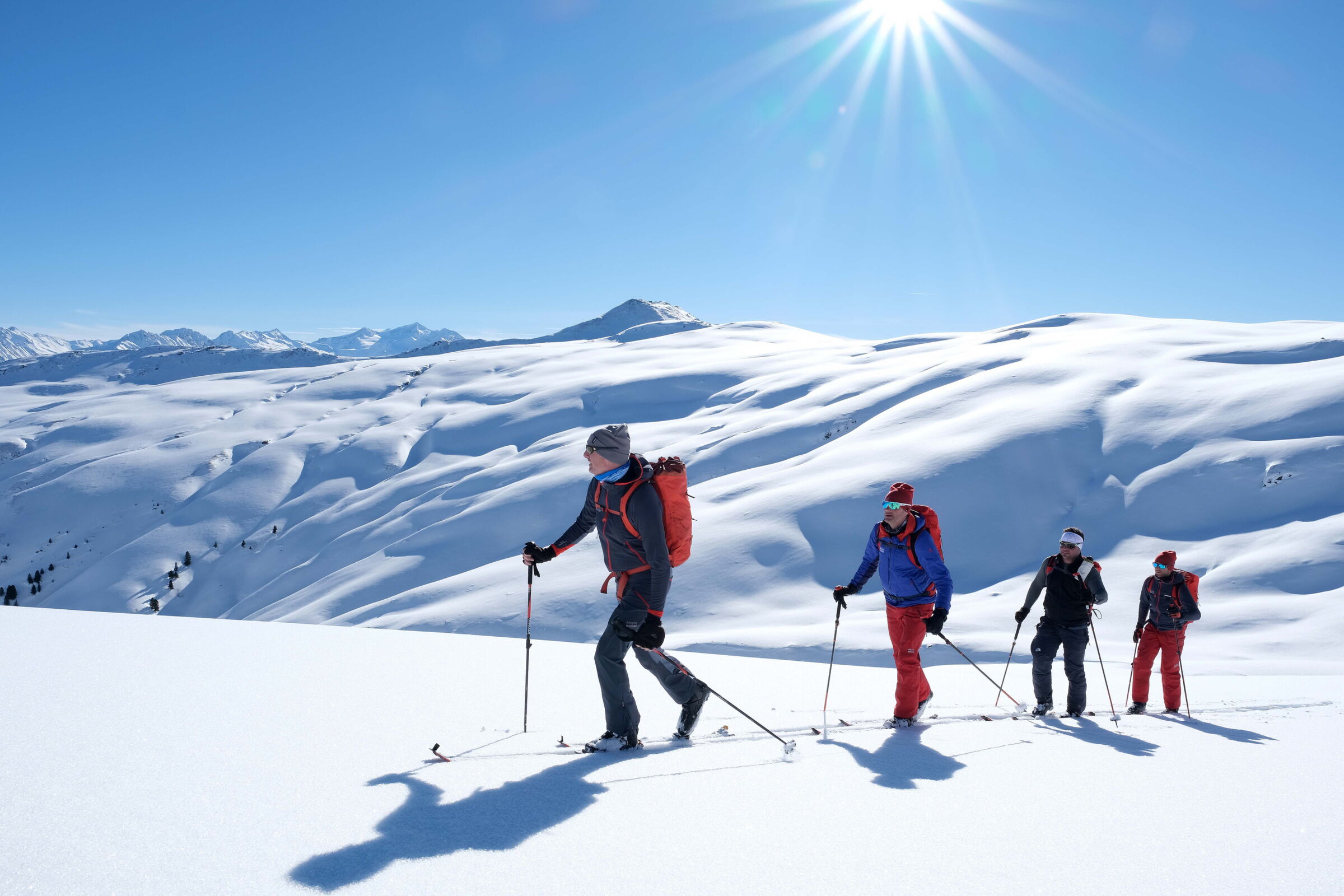Vier Personen wandern in sonnenbeschienener Schneelandschaft unter klarem blauen Himmel.