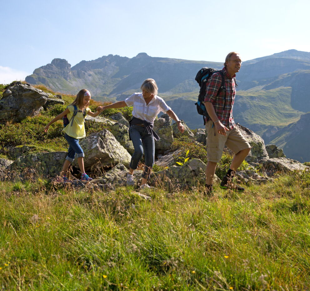 Familie wandert über grüne Wiese mit beeindruckendem Bergpanorama im Hintergrund.