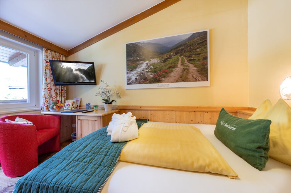 Helles Zimmer mit rotem Sessel, Holzmöbeln und beeindruckendem Bergbild an der Wand.