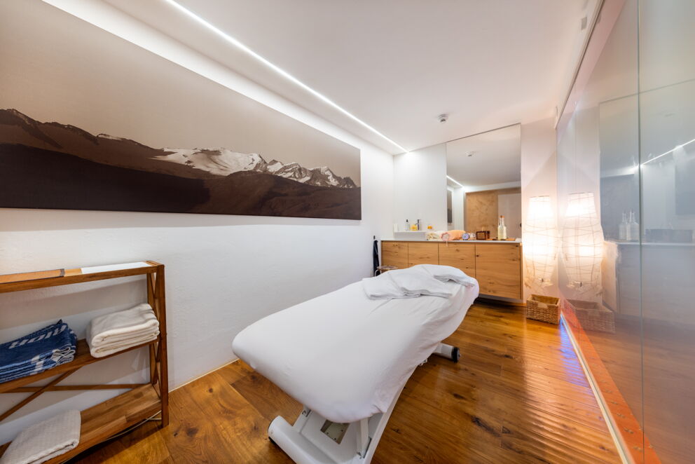 Eleganter Raum mit Massageliege, Holzregal, großem Bergbild und weichen Tüchern.