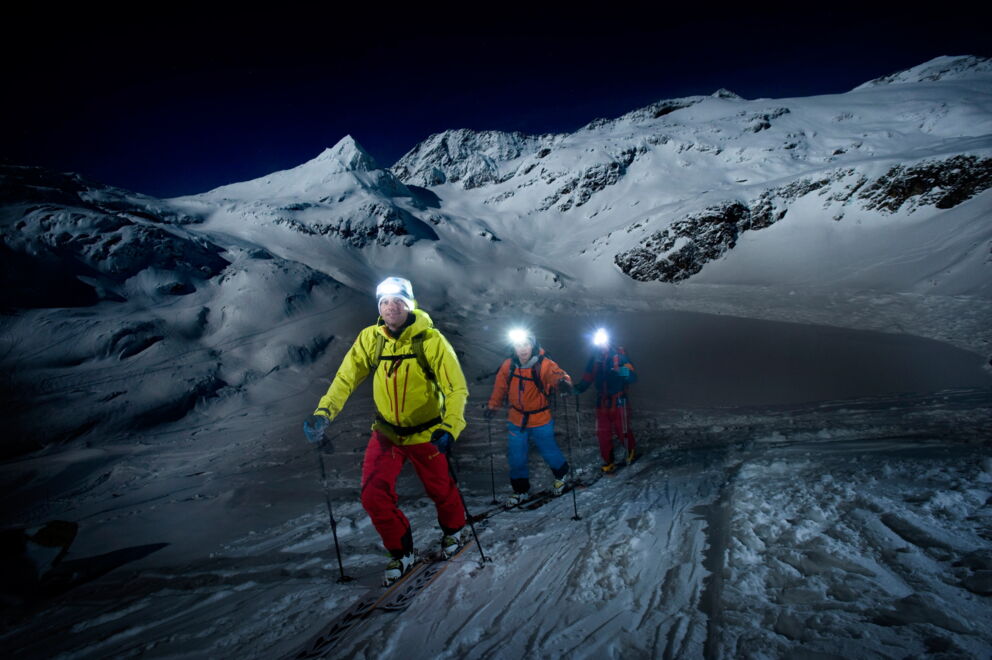 Drei Personen beim nächtlichen Aufstieg in verschneiter Berglandschaft mit Stirnlampen.
