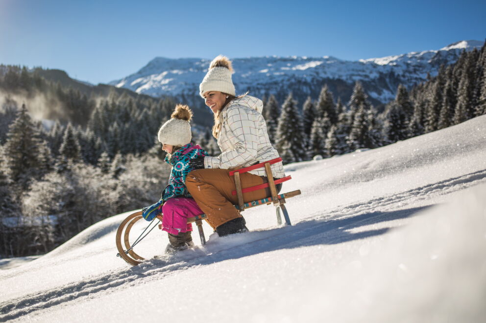 Mutter und Kind rodeln fröhlich im verschneiten Gebirge bei strahlendem Sonnenschein.