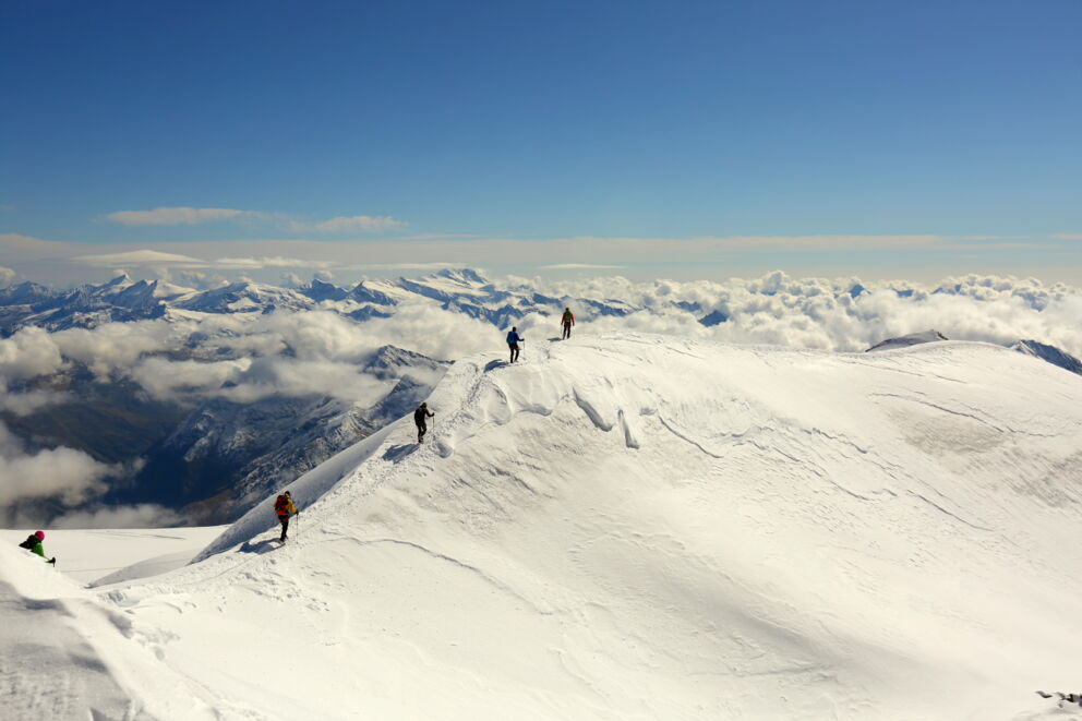 Vier Wanderer erklimmen schneebedeckten Berg unter strahlend blauem Himmel.