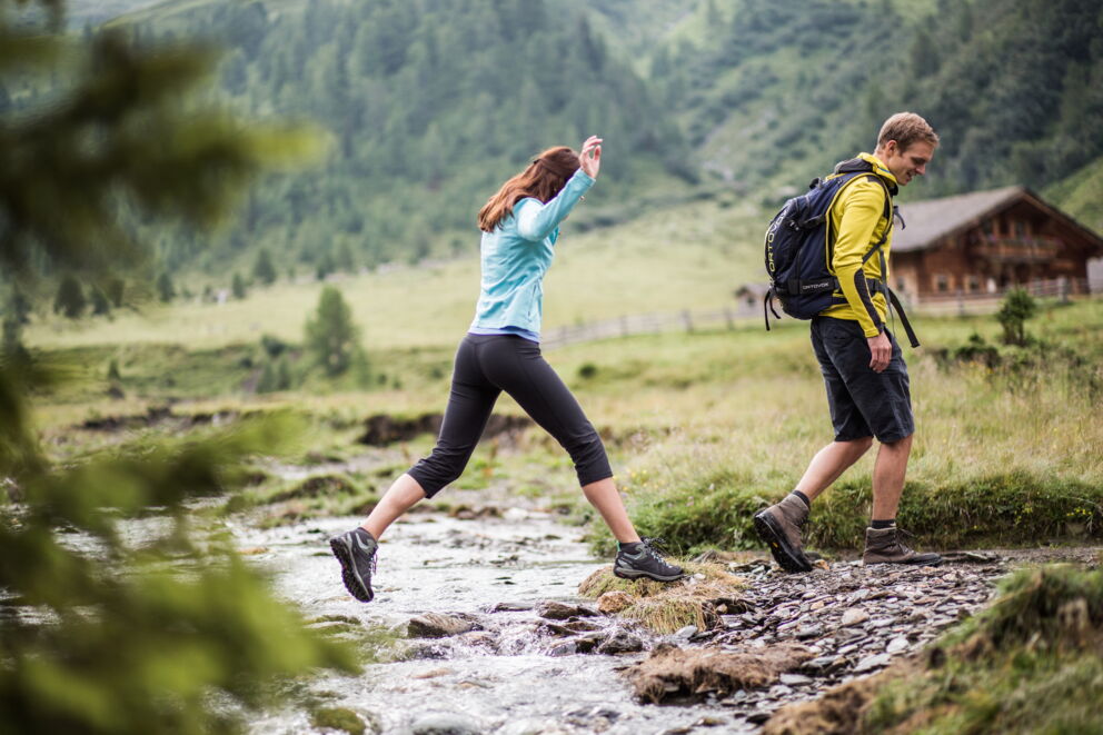 Zwei Wanderer überqueren einen kleinen Bach in alpiner Landschaft.