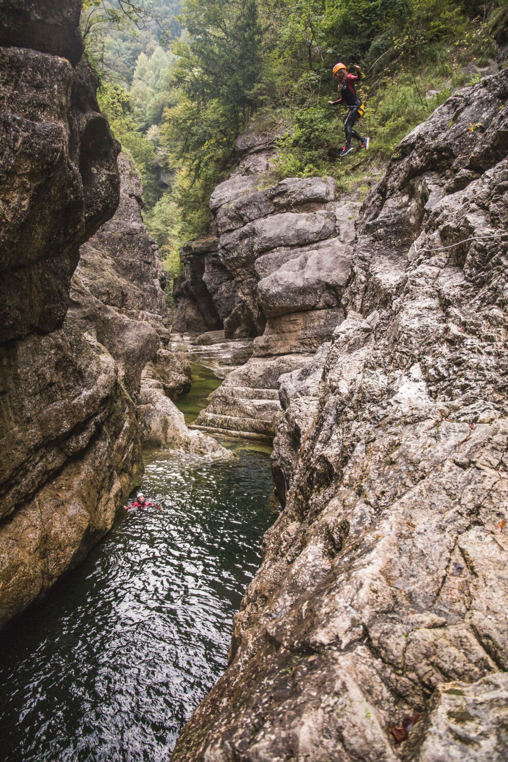Person springt in eine malerische, von Felsen umgebene Schlucht mit klarem Wasser.