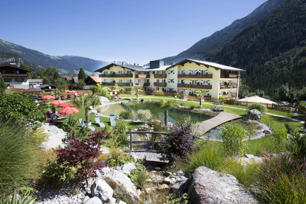 Hotel mit großem Garten, Bio-Schwimmteich und Bergblick unter strahlend blauem Himmel.