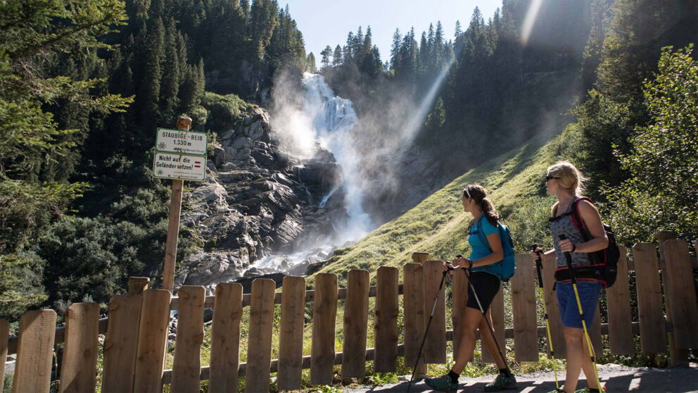 Two hikers admire a majestic waterfall surrounded by green slopes and forests.