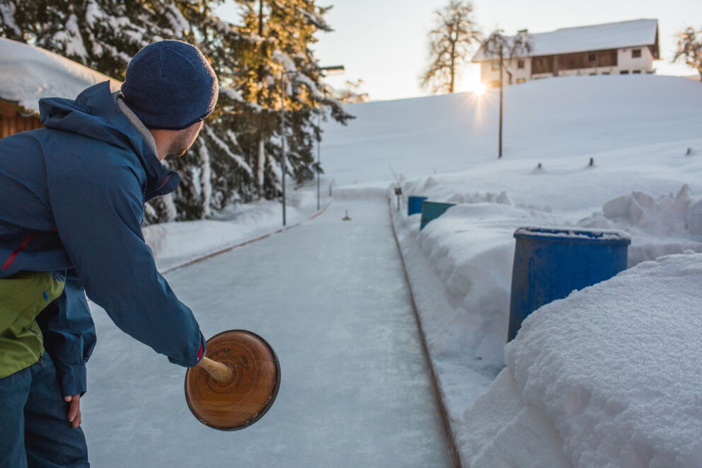 Person spielt Eisstockschießen auf schneebedeckter Bahn, Sonne im Hintergrund.