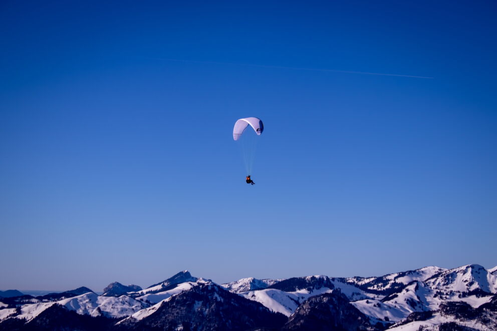 Paraglider schwebt hoch über verschneiten Berggipfeln unter klarem blauen Himmel.
