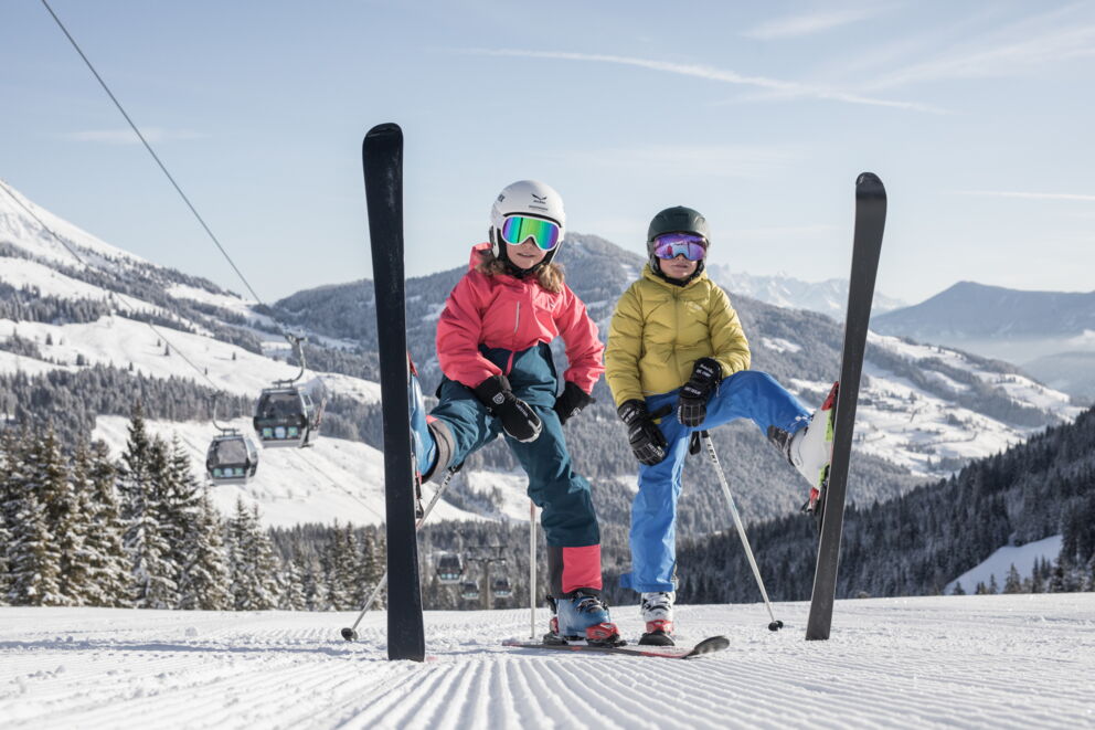 Zwei Kinder posieren mit Skiern auf schneebedeckter Piste vor Bergpanorama.
