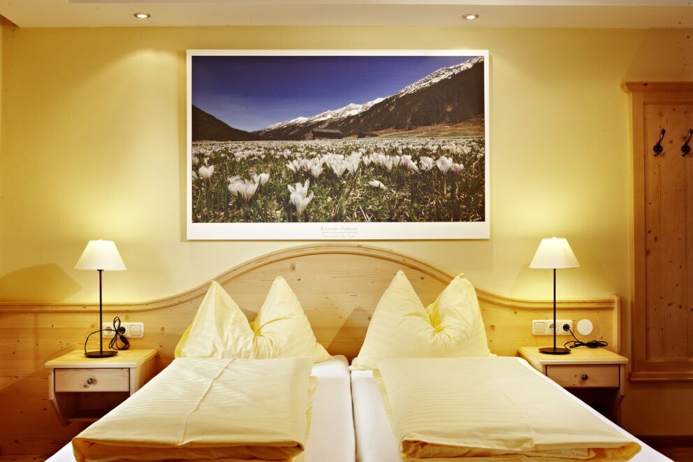 Ein Doppelbett, von vorne fotografiert, mit zwei Nachttischlampen und einem Gemälde einer blühenden Landschaft am Kopfende.