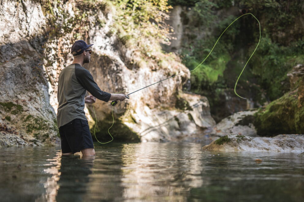 Person angelt in klarem Wasser umgeben von moosbewachsenen Felsen und Bäumen.