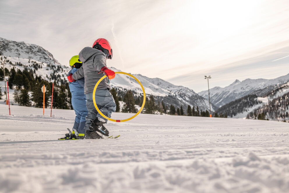 Zwei Kinder auf Skiern mit Reifen, umgeben von verschneiten Bergen und blauem Himmel.