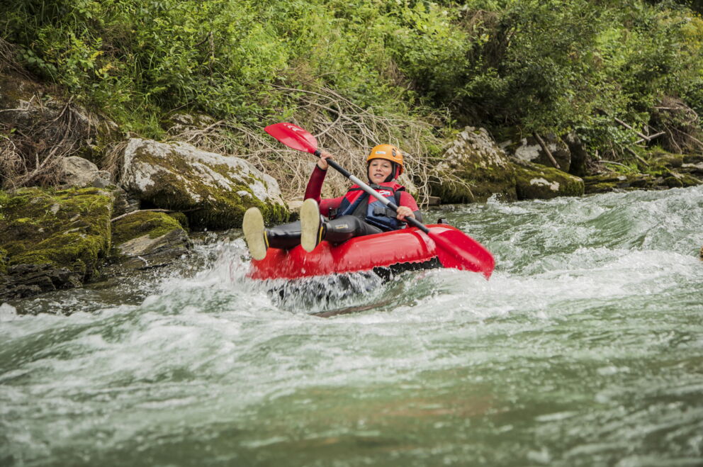 Person im Kajak auf sprudelndem Fluss, umgeben von grüner Natur.