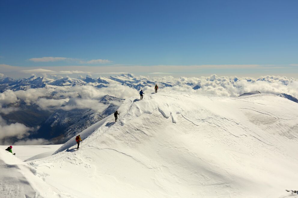 Vier Wanderer erklimmen schneebedeckten Berg unter strahlend blauem Himmel.