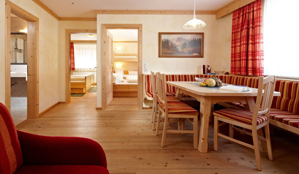 Warme Holzmöbel, roter Stoff, helle Fenster, einladender Essbereich, angrenzendes Schlafzimmer.