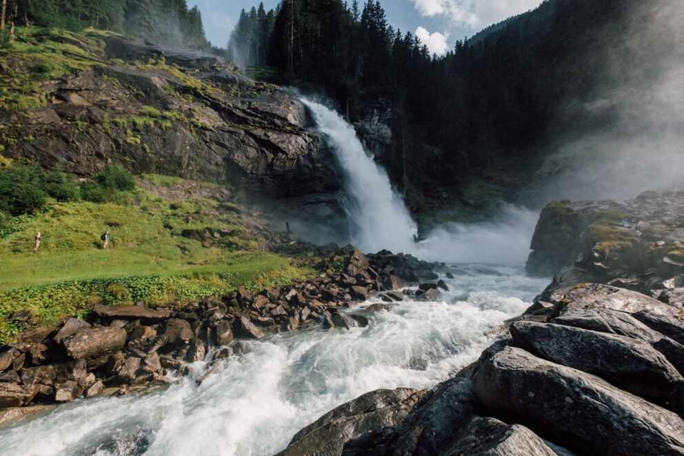 Thundering waterfalls amidst green forests and rocky landscapes, under a blue sky.