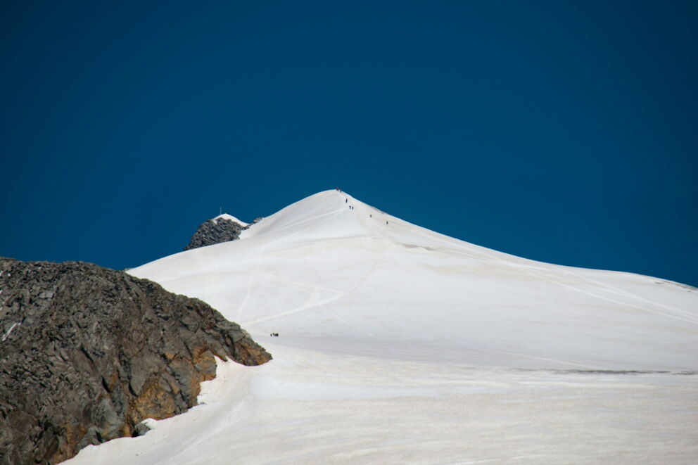 Snow-covered mountains under a bright blue sky with hikers on the summit.