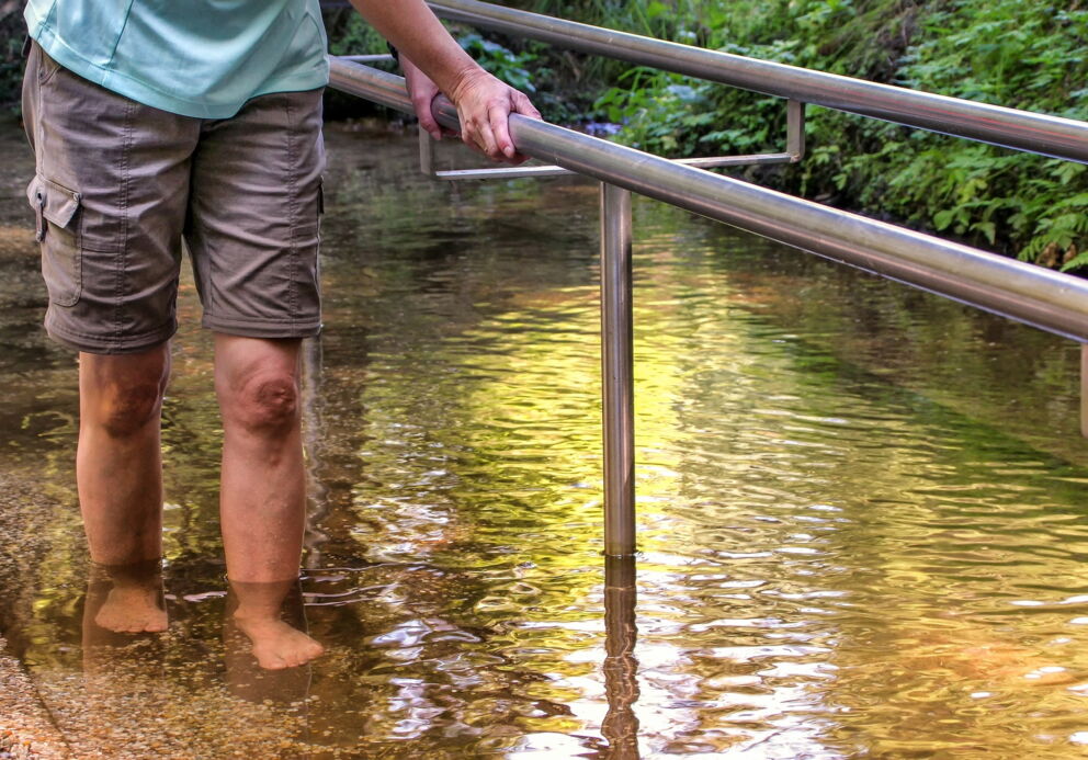 Person watet barfuß durch flaches, klares Wasser mit Handlauf.