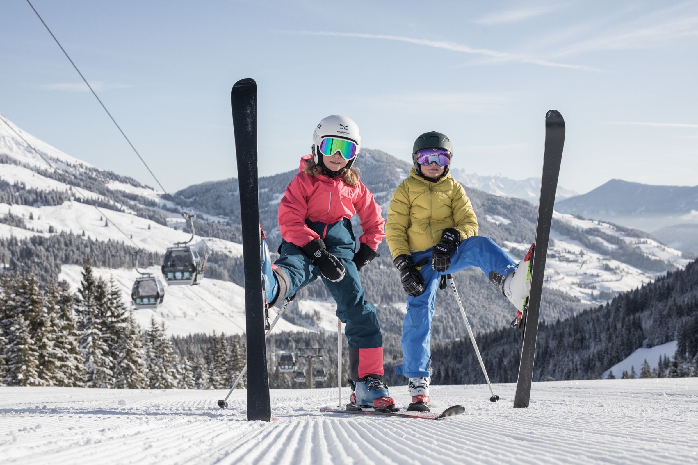 Zwei Kinder posieren mit Skiern auf schneebedeckter Piste vor Bergpanorama.